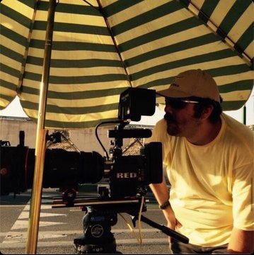 A behind-the-scenes photo of a cinematographer wearing a yellow t-shirt, cap, and sunglasses operating a RED cinema camera under a striped canopy at an outdoor production set.