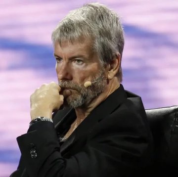 The image shows a man wearing a black suit jacket sitting in a chair with his hands placed on his lap. The background features a purple wall that contrasts with the gray hair of the man, who appears to be deep in thought or contemplation. The caption "LATEST : Strategy is facing renewed scrutiny after its massive Bitcoin holdings fell into deep unrealized losses following the latest cryptocurrency market decline". This suggests that the man might be discussing the recent events surrounding Bitcoin and the cryptocurrency market, possibly reflecting on the impact of these developments on the company's strategy.