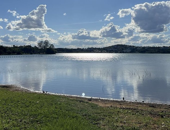 A scenic photograph of a calm lake reflecting sunlight, with green grass in the foreground and fluffy clouds scattered across a blue sky.