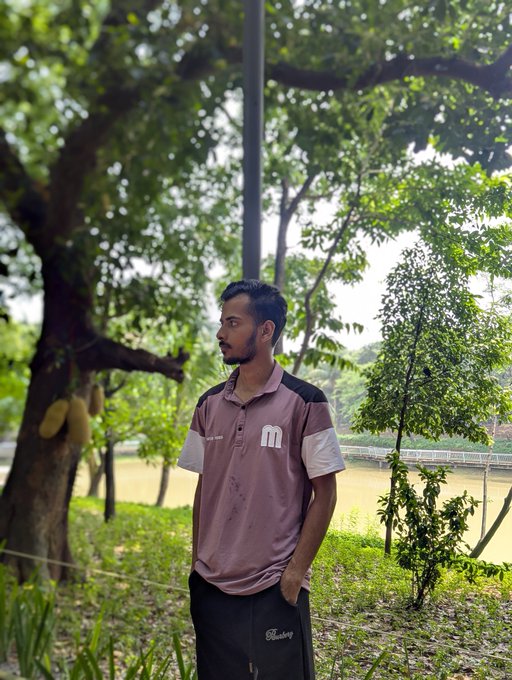 iced tea with a straw, a man in a pink shirt and black pants stands near a tree in a park, backed by a river and lush greenery.