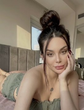A woman with dark hair styled in a high bun, wearing a green strapless top, poses on a bed with her hand resting on her cheek.