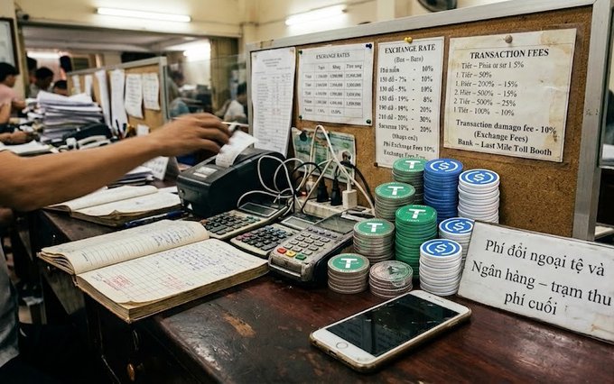 A currency exchange booth displaying rates and transaction fees, with stacks of casino chips, a calculator, and a sign in Vietnamese reading 'Phí đổi ngoại tệ và Ngân hàng - trạm thu phí cuối'.