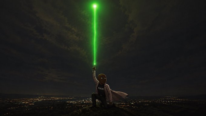 A masked figure in a white lab coat kneels atop a hill overlooking a city at night, wielding a glowing green lightsaber-style beam. The figure's shirt displays "$BLAKJAK," tying the dramatic sci-fi imagery to the cryptocurrency/brand referenced in the post.