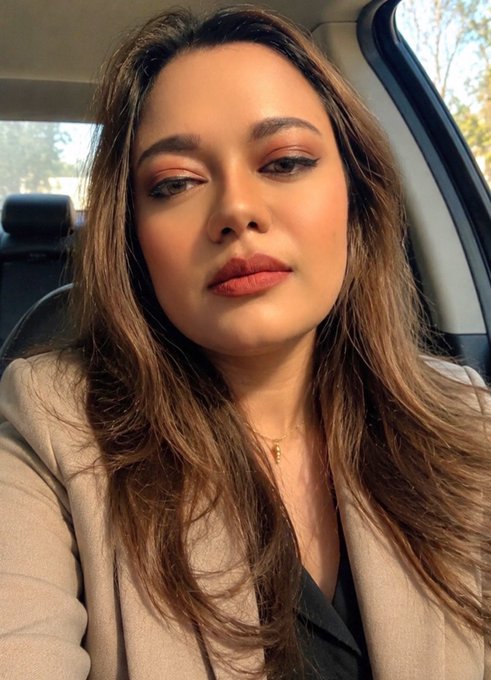A selfie of a woman with long brown hair, makeup, and a beige blazer sitting in a car.