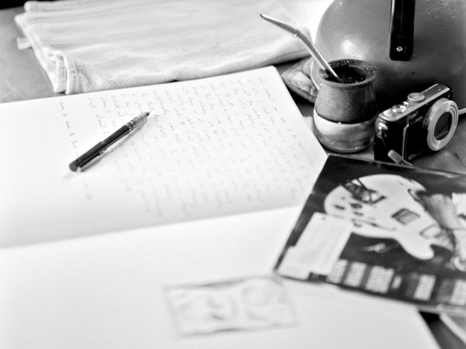 A black and white overhead shot of a desk with handwritten notes, a pen, a mate gourd with bombilla, a camera, and printed photos.