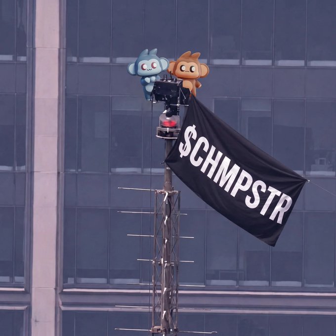 Two plush monkey characters (one blue, one orange) mounted on an antenna with a black flag displaying the text '$CHMPSTR', set against a building facade.