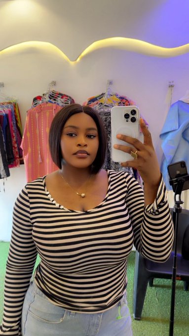 A woman in a black and white striped long-sleeve top takes a mirror selfie in what appears to be a clothing boutique or showroom, with colorful garments displayed on racks behind her. The image connects to her post as a confident mirror selfie from a stylish setting that complements her identity as a businesswoman and content creator.