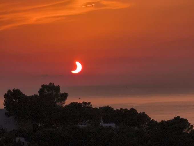 A partial solar eclipse viewed at sunset, with the crescent-shaped sun glowing orange-red above a silhouetted treeline and ocean horizon.