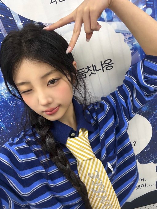 A young woman winks while posing, wearing a blue striped polo shirt with a yellow tie, making a peace sign above her head. Korean text and speech bubbles appear in the background.