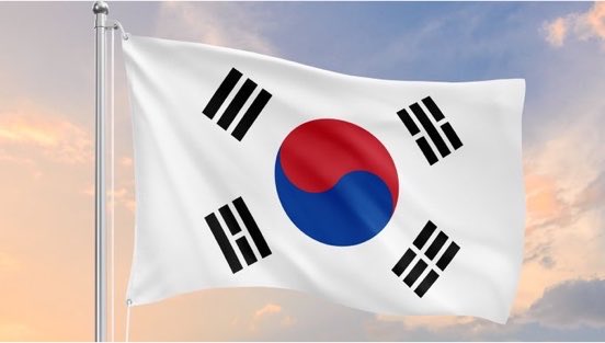 The South Korean flag, featuring its iconic red and blue taegeuk symbol and four black trigrams, waves against a sunset sky. The image symbolizes South Korea's involvement in the news about KB Kookmin Bank partnering with JPMorgan's blockchain technology for crypto-asset-style payments.