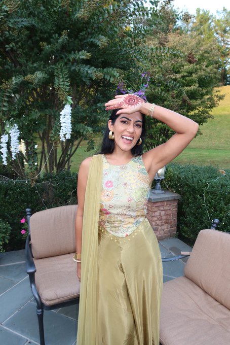 A smiling young woman in a floral and gold traditional outfit stands outdoors in a garden setting, showing off henna designs on her raised hand.
