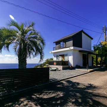 A modern two-story white house with a palm tree in the foreground, overlooking the ocean under a clear blue sky.