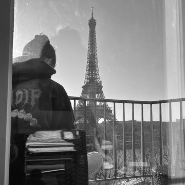 A black and white photo showing a reflection of a person on a window with the Eiffel Tower visible in the background in Paris.