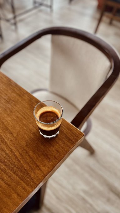 A small glass of espresso with a lemon slice garnish sits on a wooden table edge, with a modern chair and light wood floor in the blurred background.