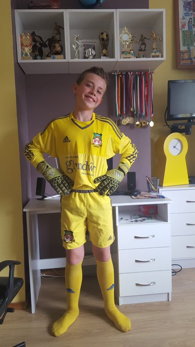 A young boy beams proudly in a full yellow goalkeeper kit featuring the "Gündüz" sponsor and Adidas branding, standing in his bedroom surrounded by shelves of trophies and medals. The image captures the "how it's going" moment, showcasing the rewards of his dedication to soccer with the gleaming awards displayed behind him serving as evidence of his journey and achievements.