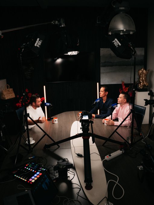Two men sit across from each other at a wooden table in a professional podcast studio, engaged in conversation with microphones set up in front of them, surrounded by professional lighting equipment and camera gear. The setup visually supports the post about a podcast interview, capturing the behind-the-scenes production environment as the two guests share perspectives from different investing worlds.