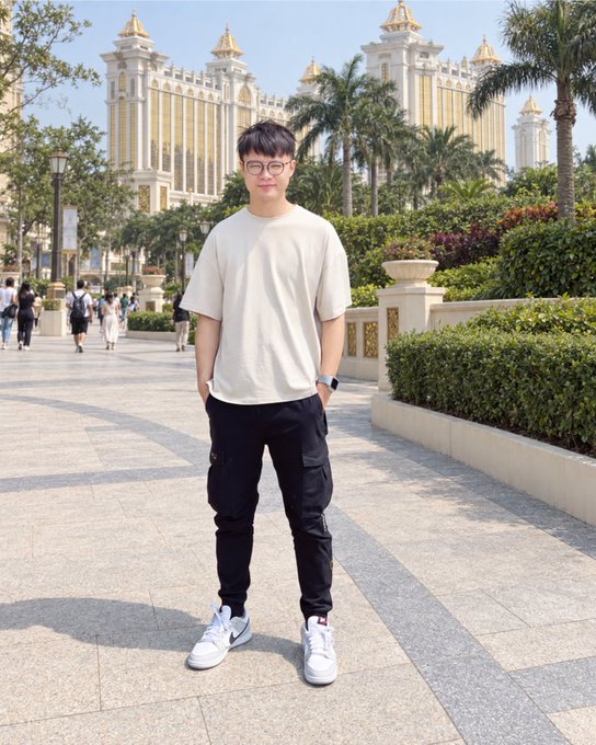 A full-body photograph of a young man with glasses wearing a beige t-shirt, black joggers, and white sneakers, standing on a paved walkway in front of an ornate golden building (resembling Galaxy Macau) with palm trees and other pedestrians in the background.