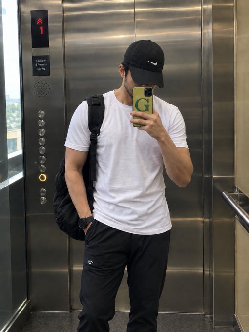 A man wearing a black Nike cap, white t-shirt, and backpack takes a mirror selfie in an elevator using a phone with a green 'G' case.