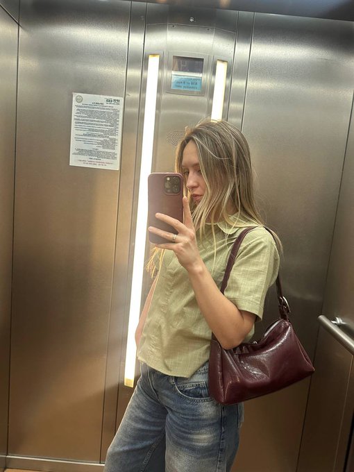 A woman takes a mirror selfie in an elevator, wearing a green button-up shirt, jeans, and carrying a burgundy handbag.