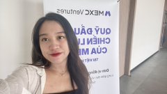 A selfie of a young woman with long dark hair standing in front of a banner with text including 'MEXEO Century' and 'YOU CAN ACHIEVE' (appearing mirrored).