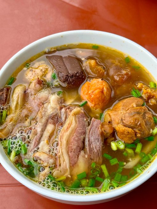 A bowl of Vietnamese soup containing broth, pork slices, meatballs, and green onions on a red table surface.