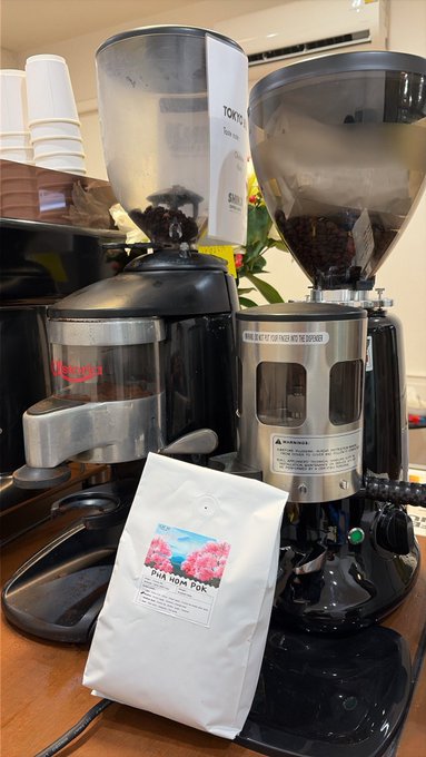 Two commercial coffee grinders filled with beans sit on a counter, with a white bag of 'Finca Roma PoK' coffee placed in front of them.