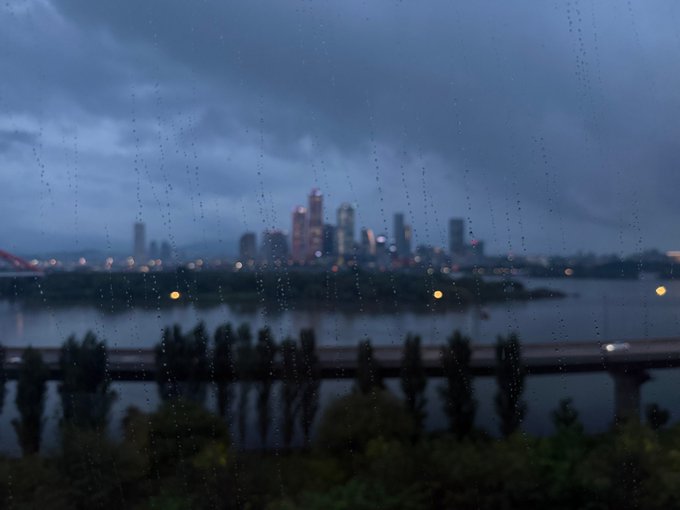 A moody cityscape at dusk, viewed through a rain-streaked window, shows a distant skyline across a river with a bridge and silhouetted trees in the foreground. The rainy, dim atmosphere mirrors the heartfelt post's message about pushing through a tough day while missing someone, the gloomy weather echoing feelings of longing and determination.