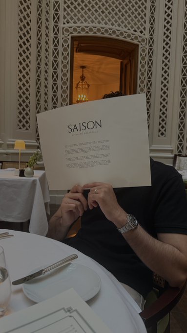 A man holds up a menu for "SAISON by Michael Tusk," covering his face while seated at an elegantly set table in a fine dining restaurant. The caption "POV: Life after you trade time based ranges" humorously suggests that mastering time-based trading strategies has afforded him the luxury of indulging in high-end dining experiences.