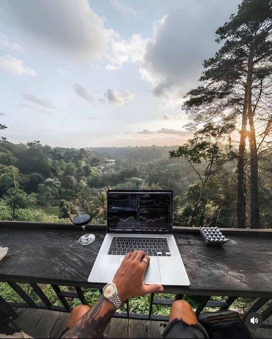 A person's tattooed hand rests on the trackpad of a MacBook displaying trading charts, with a glass of red wine beside it, overlooking a lush green valley at sunset from a balcony.