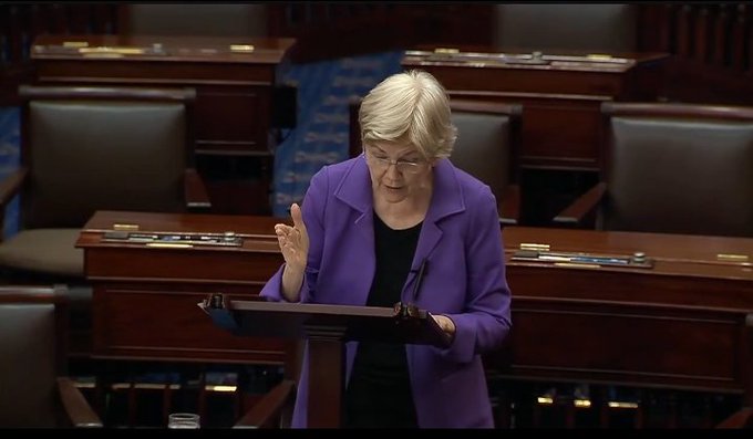 Senator Elizabeth Warren, wearing a purple blazer and glasses, stands at a podium in the Senate chamber addressing the Senate while referencing notes. The image captures her mid-speech as she publicly challenges both the cryptocurrency industry and political opponents, embodying the confrontational moment described in the caption.