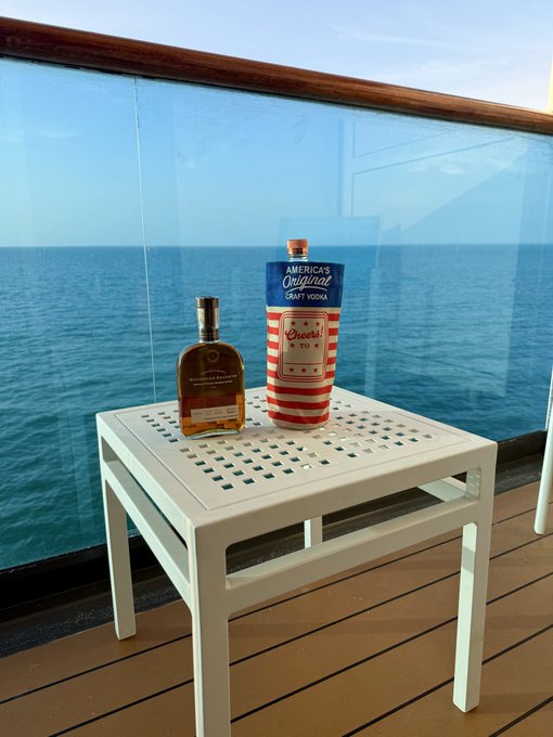 A bottle of Woodford Reserve bourbon and a patriotic red, white, and blue 'America's Original Craft Vodka' container sit on a white table on a balcony overlooking the ocean.