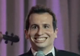 The image features a man wearing a suit, standing next to a purple curtain with a microphone attached to his neck. The man has a big smile on his face as he looks directly at the camera. There are also other people visible in the background, but they are not the main focus of the image. The post that accompanies this image is about a bean bag chair located in an office space called "SolanaSkyline".
