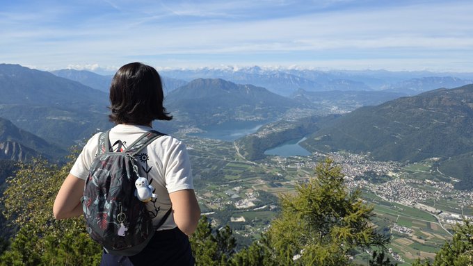 A scenic mountain landscape photo showing a person with short dark hair wearing a backpack, looking out over a valley with a river, lake, town, and snow-capped mountains in the distance.