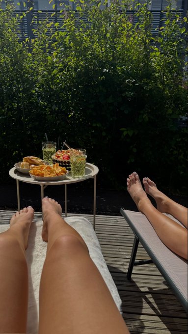 A first-person view from someone relaxing on a lounge chair on a wooden deck, with their and another person's feet visible, alongside a small table holding snacks and iced drinks, surrounded by green hedges.
