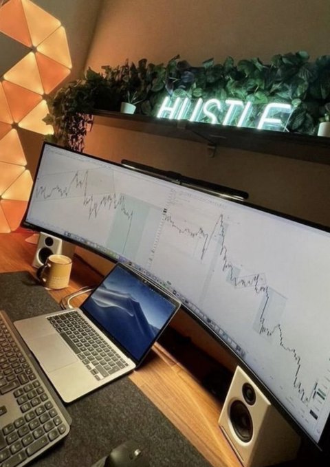 A trading setup featuring an ultrawide curved monitor displaying financial charts, a laptop, a keyboard, speakers, and a neon "HUSTLE" sign above plants.