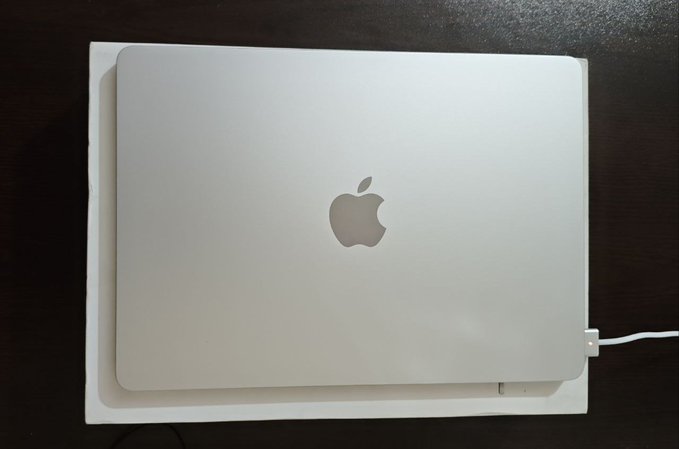 A silver Apple MacBook laptop sitting closed on top of its white packaging box on a dark wooden surface.