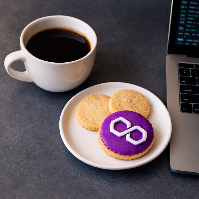 The image features a white plate with three cookies sitting next to an open laptop computer, which has a purple cookie on top of its keyboard. The background appears to be a dark gray surface. There are also two cups present in the scene - one containing coffee and another possibly filled with tea or hot chocolate.