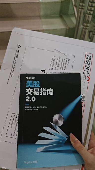 A hand holds a Bitget-branded booklet titled "美股交易指南 2.0" (US Stock Trading Guide 2.0) in front of a shipping envelope, illustrating the post about unexpectedly receiving the official Bitget guide in the mail while researching their US stock offerings.