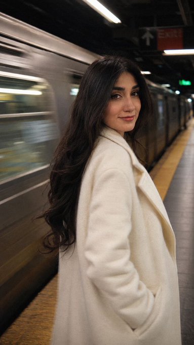 A young woman with long dark hair wearing a cream-colored coat stands on a subway platform, looking back over her shoulder as a blurred train passes behind her.