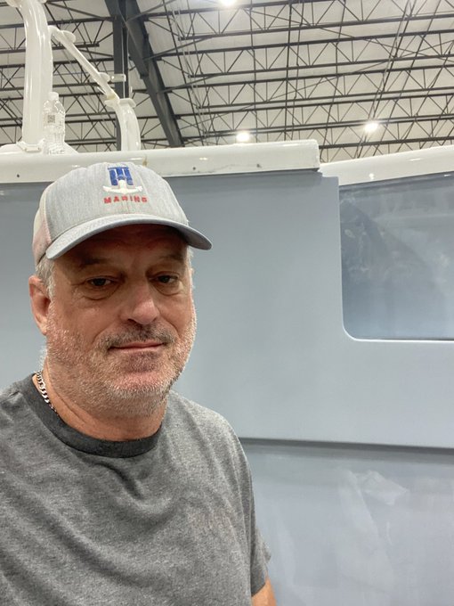 A middle-aged man wearing a gray 'H MARINE' cap and gray t-shirt takes a selfie inside what appears to be an aircraft hangar with a blue aircraft fuselage visible behind him.