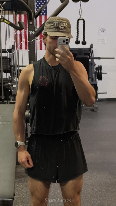 A muscular man in a black sleeveless athletic shirt, shorts, and a cap takes a mirror selfie in a gym, with "Start Time 04:11" visible on the screen.