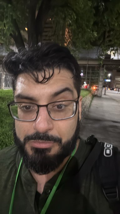A nighttime selfie of a bearded man with glasses wearing a dark green jacket and backpack strap, standing on a lit walkway with trees and city lights in the background.