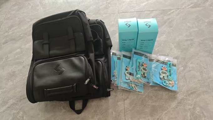 The image features a black backpack laying on its side, with several items placed inside of it. There are two blue boxes next to each other, one larger box and one smaller one. A pair of scissors can also be seen among the items. The background is gray, providing a neutral backdrop for the objects in focus. The post includes text that reads "叫你帮我拆个小箱拍个照", which translates to "I want you to take this backpack".