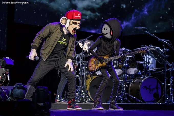Two performers on a concert stage wearing monkey costumes; one in a red 'JingleBell' cap is singing, while the other in a black hoodie is playing an electric guitar.