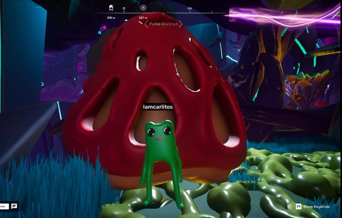 A video game scene featuring a small green frog-like character named 'lamcarilitos' standing in front of a large red mushroom in the 'Pulse District,' with 'Show Keybinds' and other HUD elements visible.
