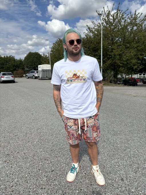 A man with teal-tipped hair and sunglasses stands in a parking lot, wearing a white t-shirt with a food graphic, patterned shorts, and white sneakers.
