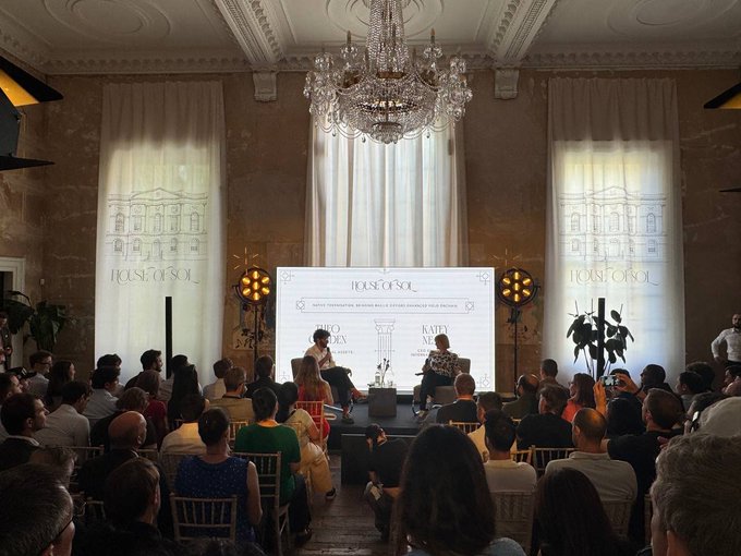 A panel discussion titled "HOUSE OF SOL" takes place on a stage in an ornate room with a grand chandelier, where speakers address an attentive audience about the Solana ecosystem in London, aligning with the post's praise for the venue as a premier meeting spot for top institutions and founders.