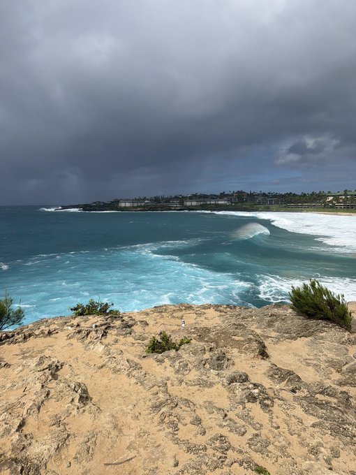 A scenic coastal view from a rocky cliff overlooking turquoise waves breaking near a shoreline with buildings, under a dramatic dark cloudy sky.