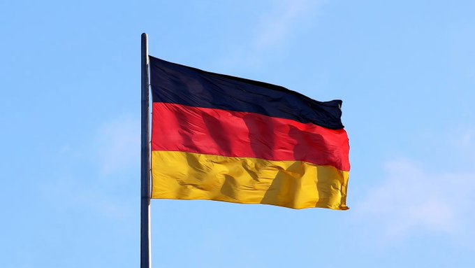 The national flag of Germany, featuring horizontal stripes of black, red, and gold, waves in the wind against a blue sky.
