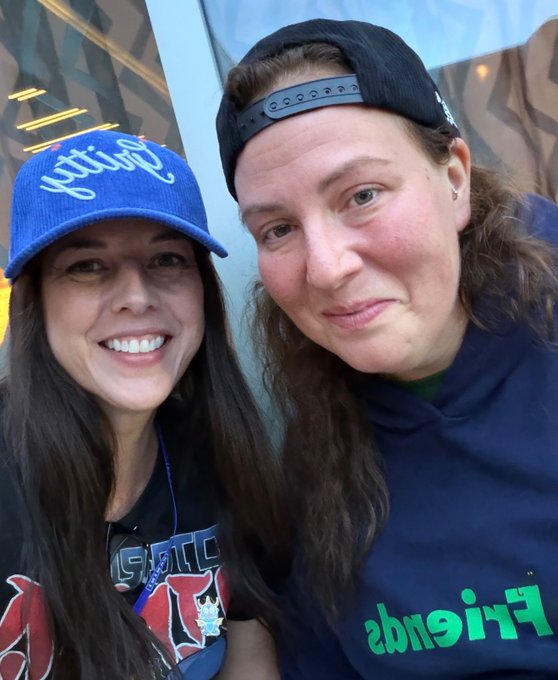 A selfie of two smiling women; the left wears a blue "Cowgirls" cap, while the right wears a black cap and a navy "Friends" hoodie.